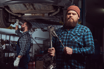Obraz na płótnie Canvas Two bearded brutal mechanics repair a car on a lift in a garage.