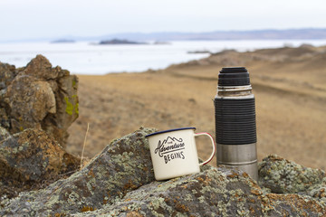 Aluminum enameled camp mug with tea and a metal thermos on the stony shore of Lake Baikal in early spring on a picturesque natural background among the stones covered with moss.