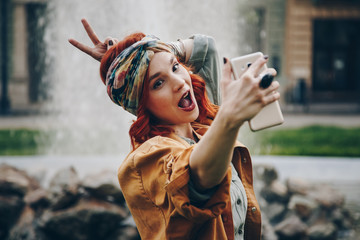 beautiful young woman taking selfie picture outdoors. hippie fashion blogger on vacation, taking self portrait with smartphone. street style, music festival portrait of authentic and fun young girl. - obrazy, fototapety, plakaty