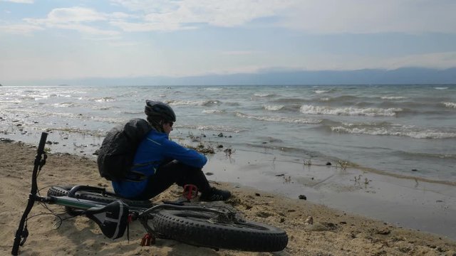 Fat bike also called fatbike or fat-tire bike in summer driving on the beach. The guy and his bike rest on the sandy beach. He looks at the sea and enjoys his sight. Throws stones at sea.