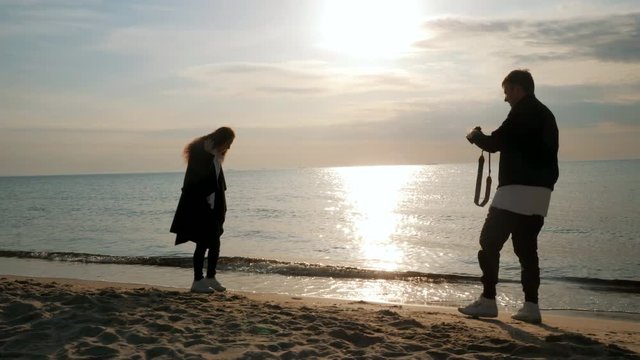 Man takes pictures of his beloved on the seashore in spring. Photoshoot of beautiful people on the beach. Happy married adult couple having fun, playing, fooling. Girl posing for guy with camera