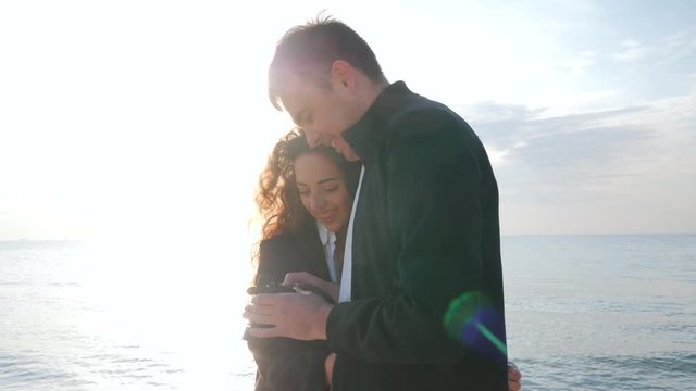 Man takes pictures of his beloved girl on the seashore in spring. Couple watching photos on the camera after a photoshoot and smiling together. 4k