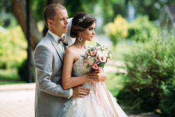 beautiful wedding couple hugging in park with green trees on background. Groom in a business gray suit, white shirt in a bow tie and a buttonhole. Bride in a luxury dress with a veil and a bouquet of