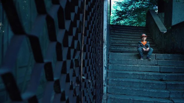 A lonely child is sitting on empty steps on the street, a 5 year old boy with a serious face posing on the camera