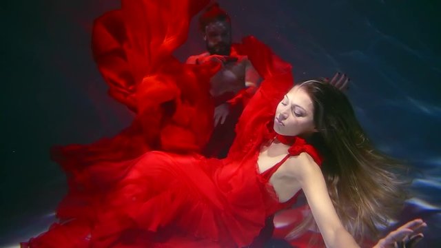 underwater shot of two people in a pool, woman is wearing red long dress is at gunpoint of anger man