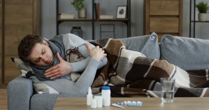 Unwell young man in a scarf and covered with a plaid caughing hard while lying on the gray couch in the cozy living room, having a flu. Indoor