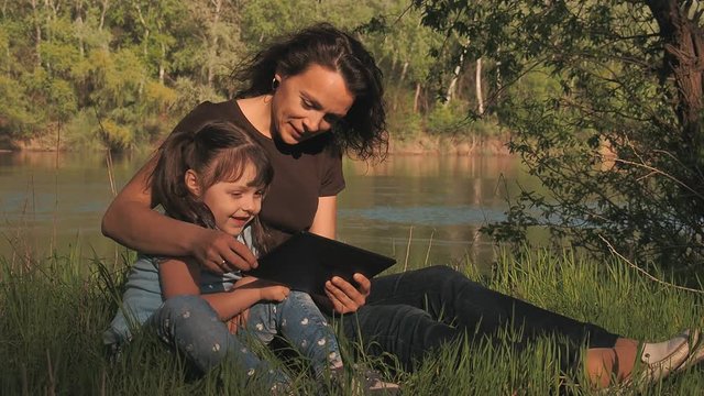 Family in nature with a tablet. Mom and daughter on the river bank with a gadget.