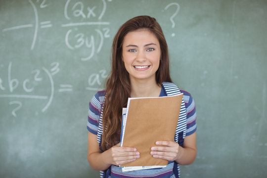 Portrait of schoolgirl holding books against chalkboard in classroom