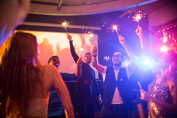 Group of trendy young people celebrating holiday in nightclub burning flaming sparklers on dance floor, copy space