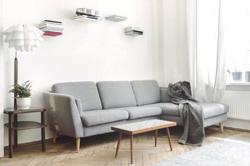 Modern interior of living room with small design table, stylish sofa and lamp. White walls with shelves from books and mock up frame. Brown wooden parquet.