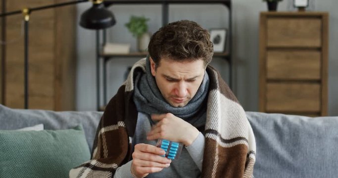 Unwell upset man in the warm scarf and plaid sitting on the couch at home, caughing hard and holding pills in a hand. Inside