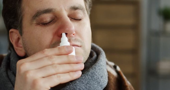 Close up of the unwell caucasian man in the warm scarf clogging up his nose with medical drops as he having a flu. At home. Inside