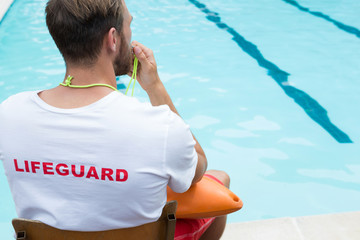 Lifeguard sitting on chair and blowing whistle at poolside - Powered by Adobe