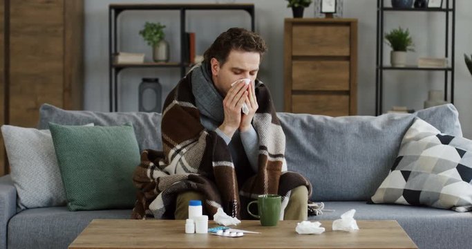 Unhealthy young man in a scarf and a plaid blowing his nose in a napkin while sitting on the sofa under the plaid in the living room, having a bad cold. Indoor