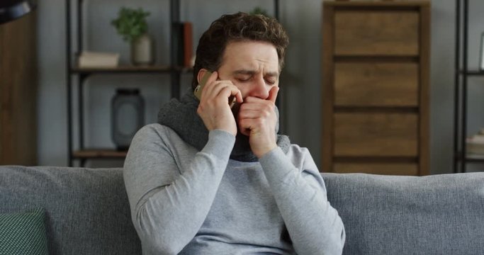 Unwell young man in a scarf and a plaid talking on the phone while sitting on the sofa, sneezing and caughing as having a cold. Inside
