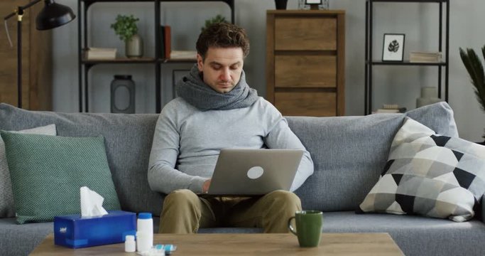 Very ill caucasian man in a gray scarf having hard caugh while typing on the laptop computer on the sofa in the living room, having a flu. Indoor