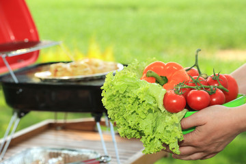 Miska z warzywami, sałata, pomidory, papryka w dłoniach kobiety i grilem w tle.