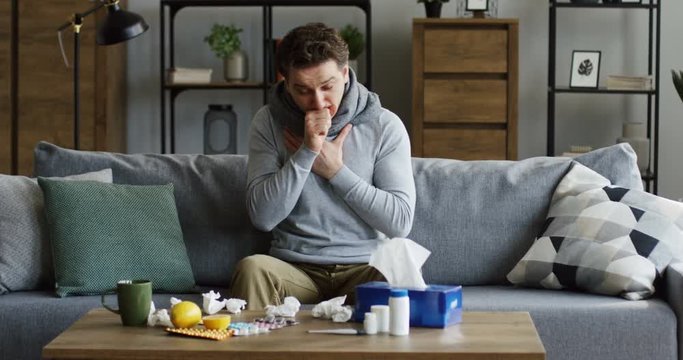 Caucasian unwell man in the warm gray scarf having a hard caugh spasm as having a cold or influenza, sitting on the couch in the cozy living room. At home. Indoor