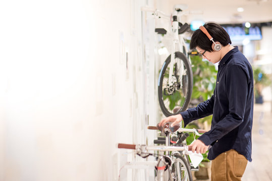 Young Asian Man With Headphones And Glasses Looking For Bicycle At Bike Shop In Department Store, Urban Lifestyle In Shopping Mall Concept