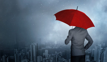 Businessman standing while holding an red umbrella over storm in city huge rain background. Business crisis and insurance concept.