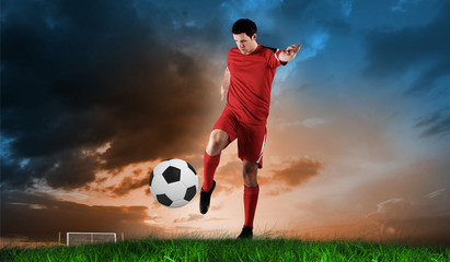 Football player in red kicking against green grass under blue and orange sky
