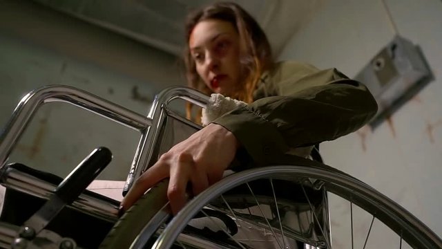 frustrated unhappy woman with bloody bruises on face is trying to rotate big wheel of gurney chair