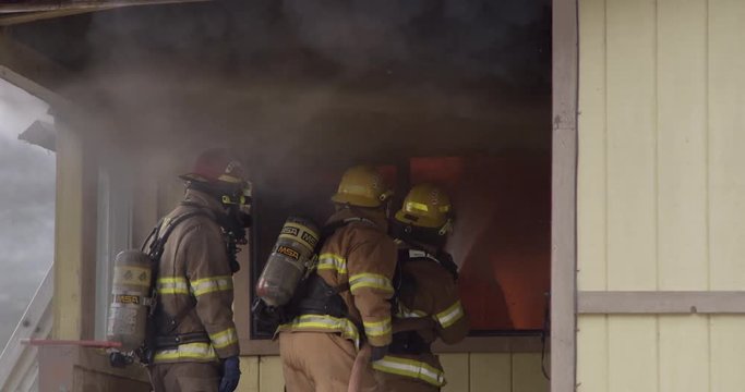 Slow-motion shot of three firemen on the porch of a house spraying water through a window