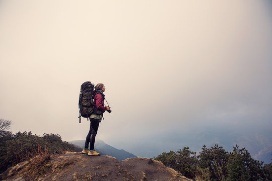 Female traveller photographer with big backpack staying on the top of mountain in jungle forest. Take pictures and enjoy view in mountains trekking, Nepal