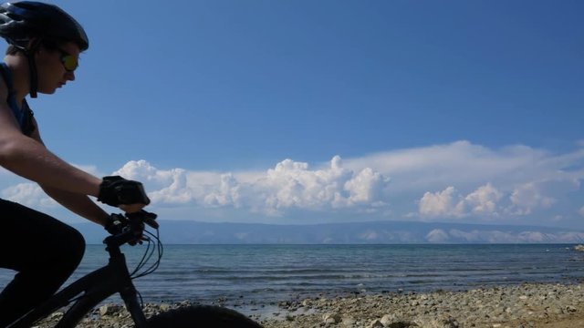 Fat bike also called fatbike or fat-tire bike in summer driving on the road. The guy rides by the hill on a sand clay path, behind him the shore by the sea. Slow motion shooting 180fps.