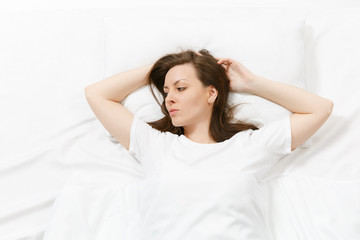 Fototapeta premium Top view of beautiful young woman lying in bed with white sheet, pillow, blanket. Calm pretty female looking aside, spending time in room. Rest, relax, good mood concept. Copy space for advertisement.