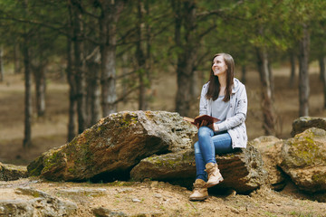 Naklejka na ściany i meble Young relaxing smiling beautiful woman in casual clothes sitting on stone studying reading book in big city park or forest on green background. Student learning, education. Lifestyle, leisure concept.
