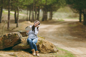 Naklejka na ściany i meble Young relaxing smiling woman in casual clothes sitting on stone listening music with headphones and tablet pc computer in city park or forest on green background. Student lifestyle, leisure concept.