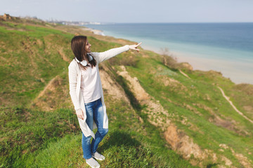 Young smiling pretty woman in light casual clothes with headphones looking aside pointing index finger on horizon standing in field near water on green background. Lifestyle, leisure concept.