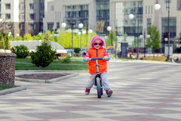 Active blond kid driving run bike the street alley of the city. Funny casual clothing vest jacket, sunglass and hood with bunny ears. Balance bike concept.