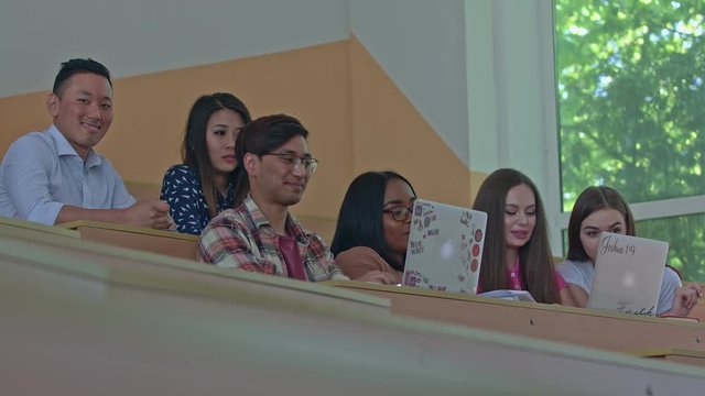 Positive students studying during lesson in the university classroom with modern desing and big, spacy panoramic windows. Working with laptops, smartphones, joking,reading, writing, being positive.