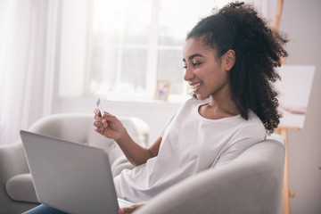 Pleasant activity. Cheerful happy woman sitting in front of the laptop with a credit card while doing online shopping