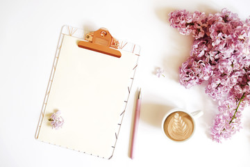 Blank paper sheet on a clipboard, cup of coffee, pink pen & lilac flowers in minimal arrangement on...