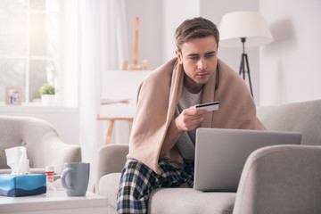 Internet pharmacy. Cheerless ill man holding his credit card while buying medicine online