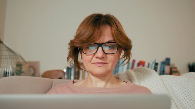Beautiful Adult Woman with Laptop is Sitting on Sofa in a Cozy Room. Screen is Reflecting in Glasses