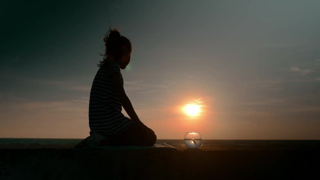 wonderful idyllic scene sitting little kid girl's silhouette and round fish tank floating fish outdoors on backdrop of the ocean at sunset in summer.