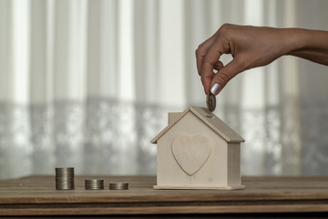 Hand putting coin in house piggy bank on wood