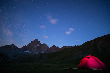 Camping under starry sky and milky way at high altitude on the Alps. Illuminated tent in the foreground and majestic mountain peak in the background. Adventure and exploration in summertime.