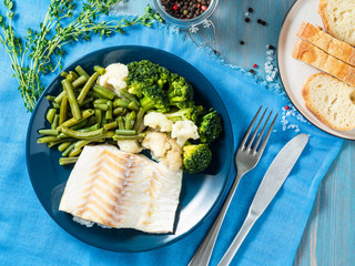 Baked sea fish cod fillet with vegetables - broccoli, green beans and cauliflower  on blue plate, bread, blue napkin, wooden background, top view. Proper diet