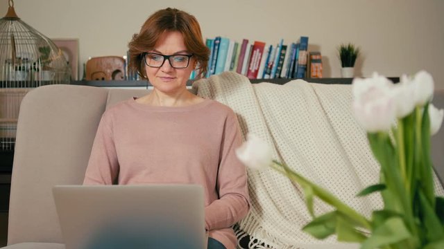 Beautiful Adult Woman with Laptop is Sitting on Sofa in a Cozy Room. Screen is Reflecting in Glasses