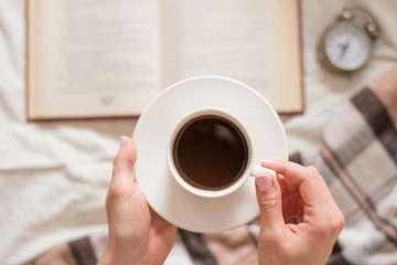 A cup of coffee in the hands of a girl with an alarm clock and a book on a brown plaid. Top view, morning concept