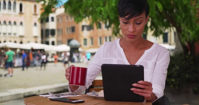 Modern black female drinks coffee while browsing the web on her tablet sitting outside at a restaurant in Venice, Attractive woman reads the news on her pad at a cafe in Venice, Italy, 4k