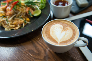 A cup of Cappuccino coffee put on wooden table with food tablet and tea in background.