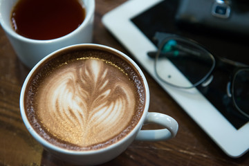 A cup of Cappuccino coffee put on wooden table with books with tablet glasses car key pen and tea in background.
