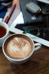 A cup of Cappuccino coffee put on wooden table with books with tablet glasses car key pen and tea in background.