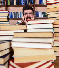 Man on nervous face between piles of books in library, bookshelves on background. Scientific discovery concept. Teacher or student with beard wears eyeglasses, sits at table with books, defocused.
